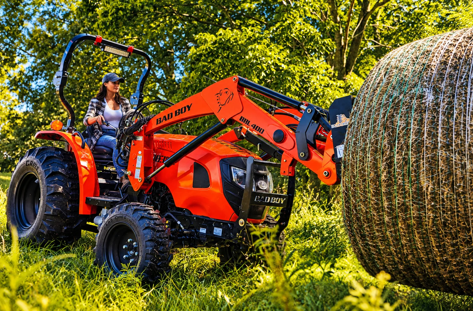 Woman operating a Bad Boy tractor carrying a hay bale in a sunlit field