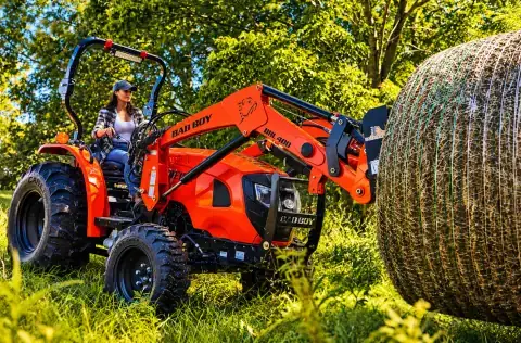 Woman operating a Bad Boy tractor carrying a hay bale in a sunlit field